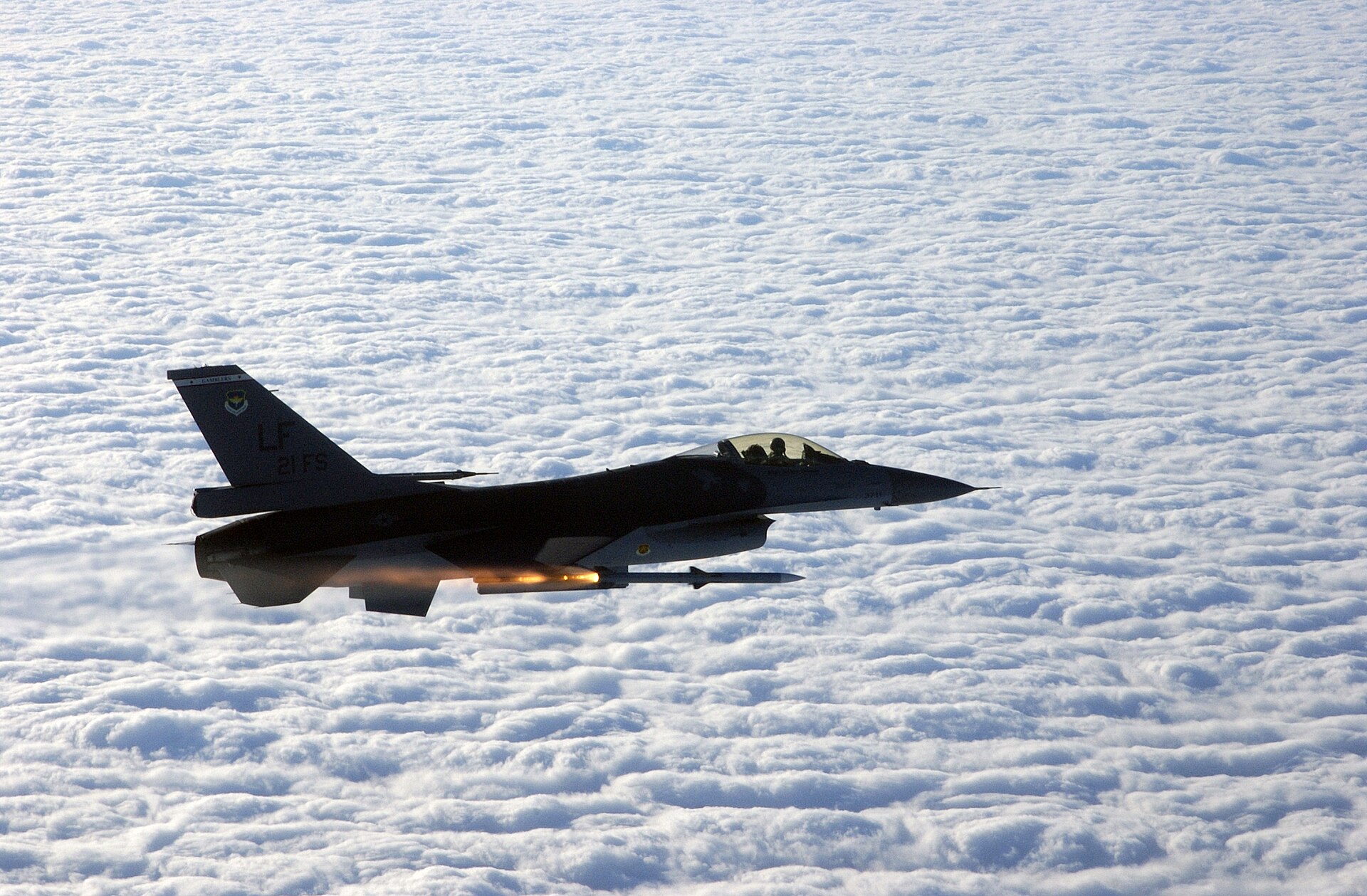 雲海上空,一架機尾漆著 LF 與 21 FS 字樣的美國空軍 F-16 從機翼下方射出一枚 AIM-120 飛彈,火箭尾焰正在點燃