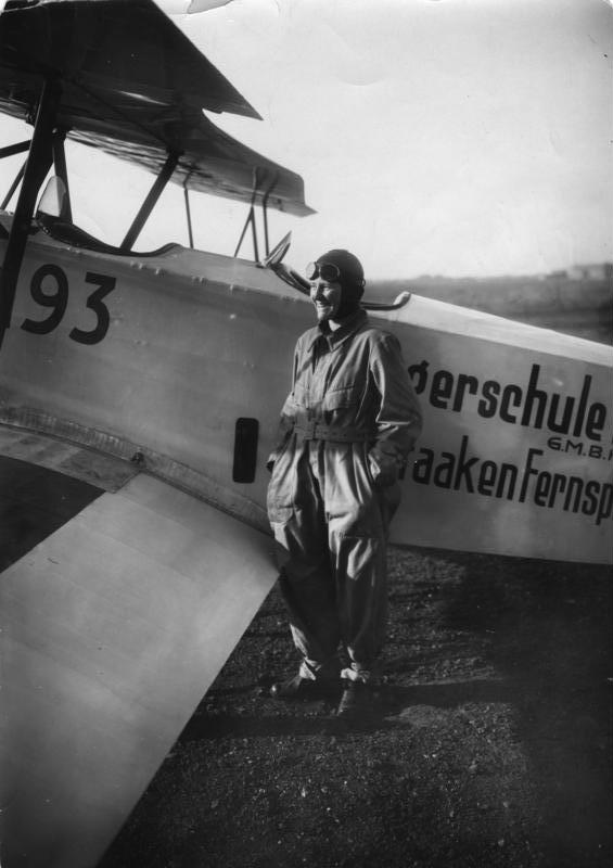 Marga von Etzdorf 穿著連身飛行服、戴飛行帽，站在一架雙翼教練機旁邊，機身漆著飛行學校的字樣