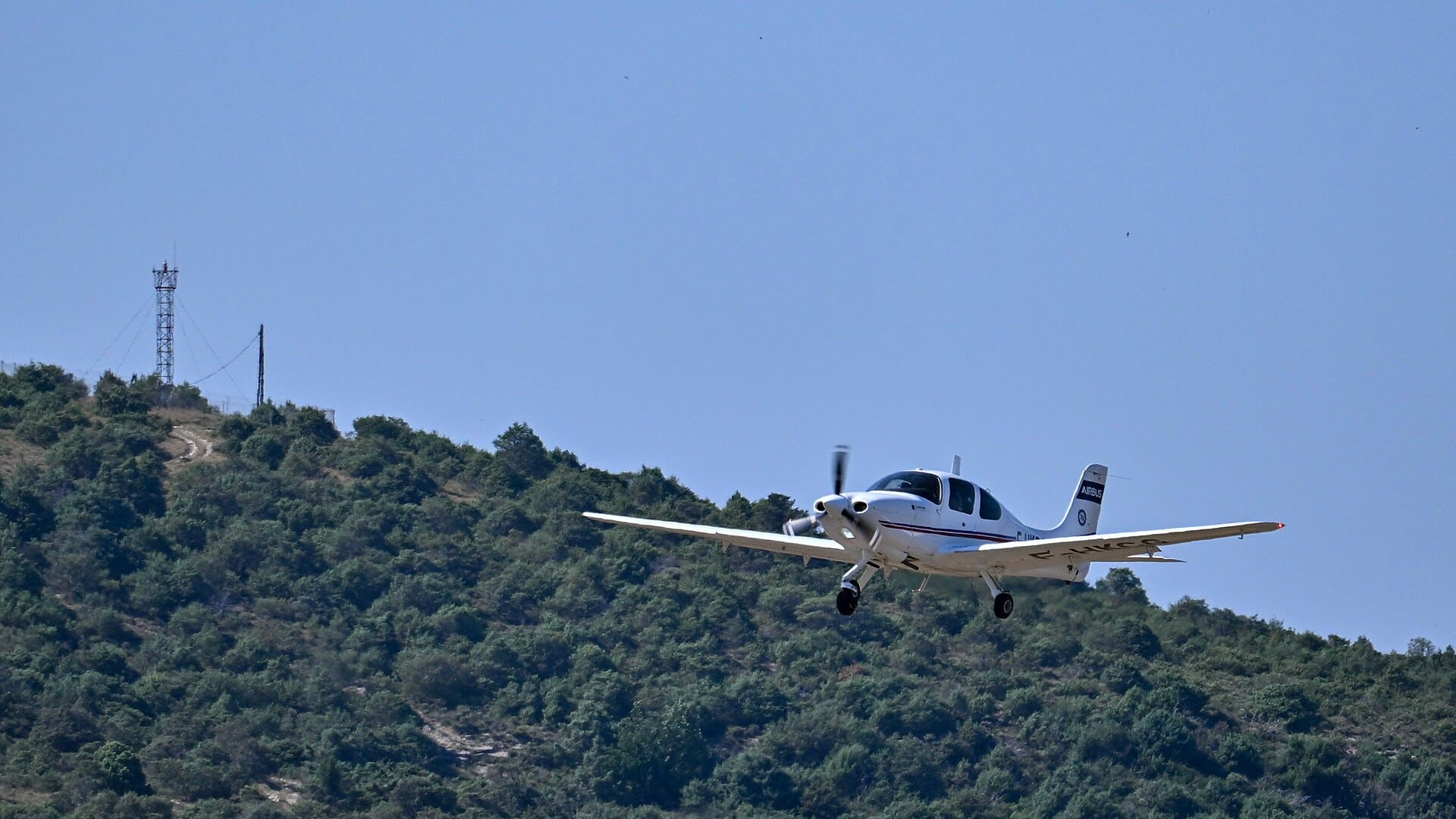 一架白色單發動機教練機 Cirrus SR20 放下起落架低空通場，機身側面有 F-HKCG 註冊編號，尾翼上是 AIRBUS 字樣，背景為長滿灌木的山丘與無雲藍天