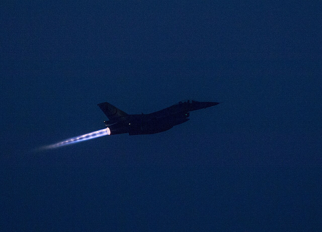 深藍色的夜空中，一架以色列空軍 F-16I 戰鬥機起飛後爬升，機身只剩深色剪影，尾管的後燃器噴出一道亮藍白色的火焰