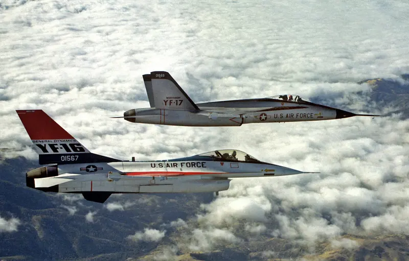 雲層上空，塗紅白藍塗裝的通用動力 YF-16 與銀色的諾斯洛普 YF-17 並排飛行，兩機翼尖都掛著響尾蛇飛彈