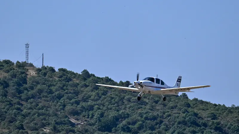 一架白色單發動機教練機 Cirrus SR20 放下起落架低空通場，機身側面有 F-HKCG 註冊編號，尾翼上是 AIRBUS 字樣，背景為長滿灌木的山丘與無雲藍天