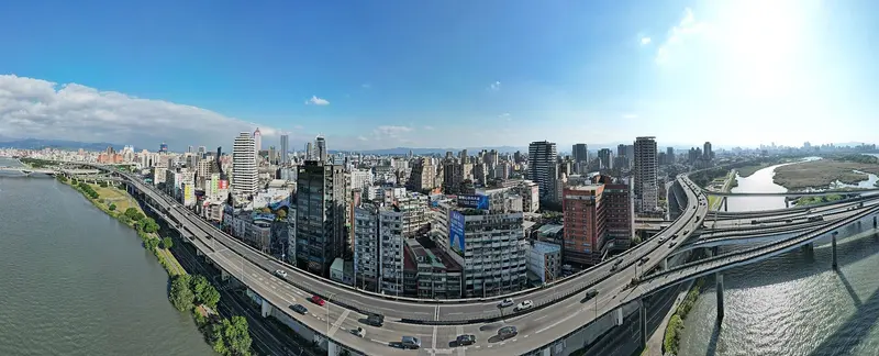 從淡水河上空俯瞰台北市西區，高架快速道路沿著河岸穿過密集的市區大樓，遠方是丘陵與市中心天際線
