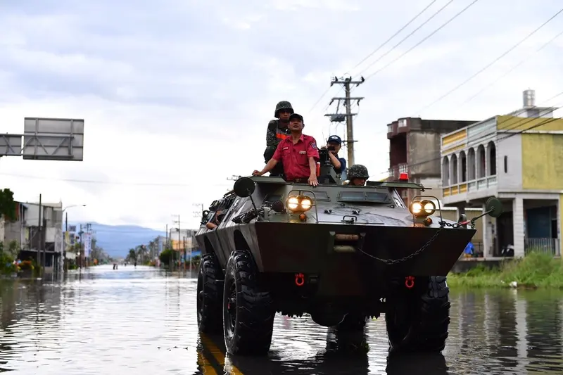 國軍裝甲車行駛在屏東淹水的街道上，車頂站著士兵與一名穿紅色制服的消防人員，兩旁街屋泡在水中