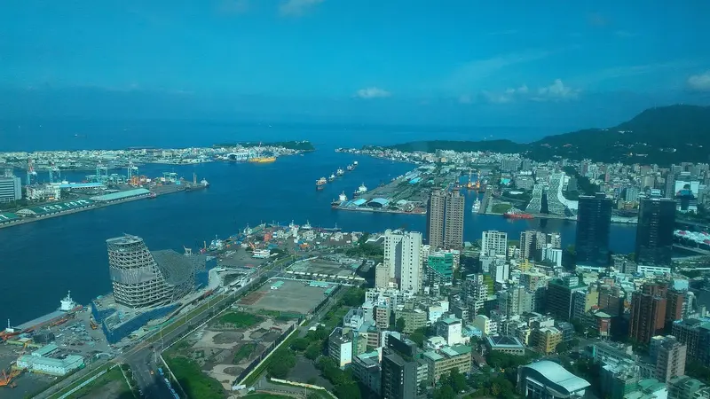 高雄港第一港口空拍，貨櫃碼頭與起重機沿著岸邊排列，貨船停靠在航道兩側，後方是高雄市區與壽山